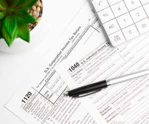 Tax forms 1040 and 1120 with a pen, calculator, and a potted plant on a white desk.