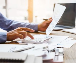 Person uses a calculator and reviews documents at a desk with a laptop and paperwork scattered around.